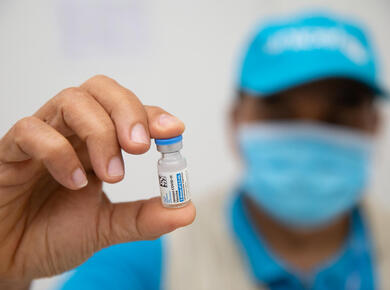 hand holding vaccine vial with blurred, masked face of health worker in background